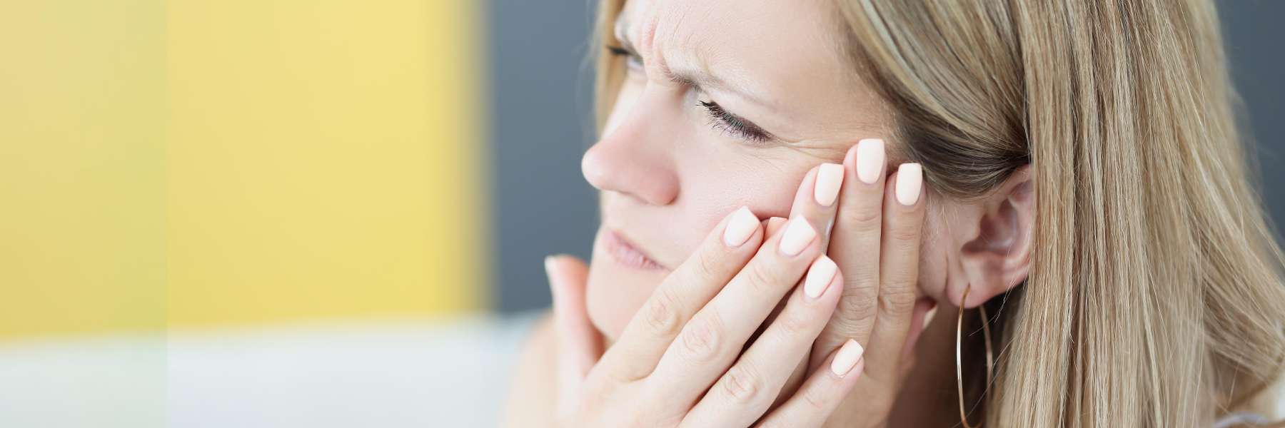A blonde woman holding her jaw with a pained expression, possibly experiencing a toothache.